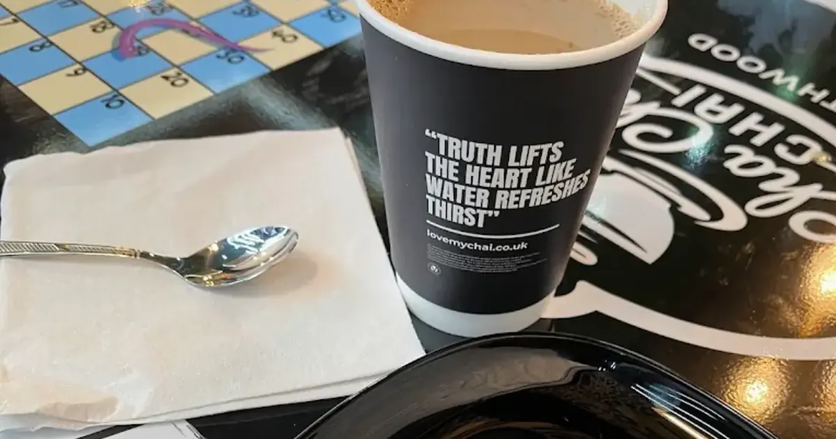 A takeaway cup of milky chai on a snakes and ladders table at Cha Cha Chai, next to a round chocolate dessert with a yellow drizzle