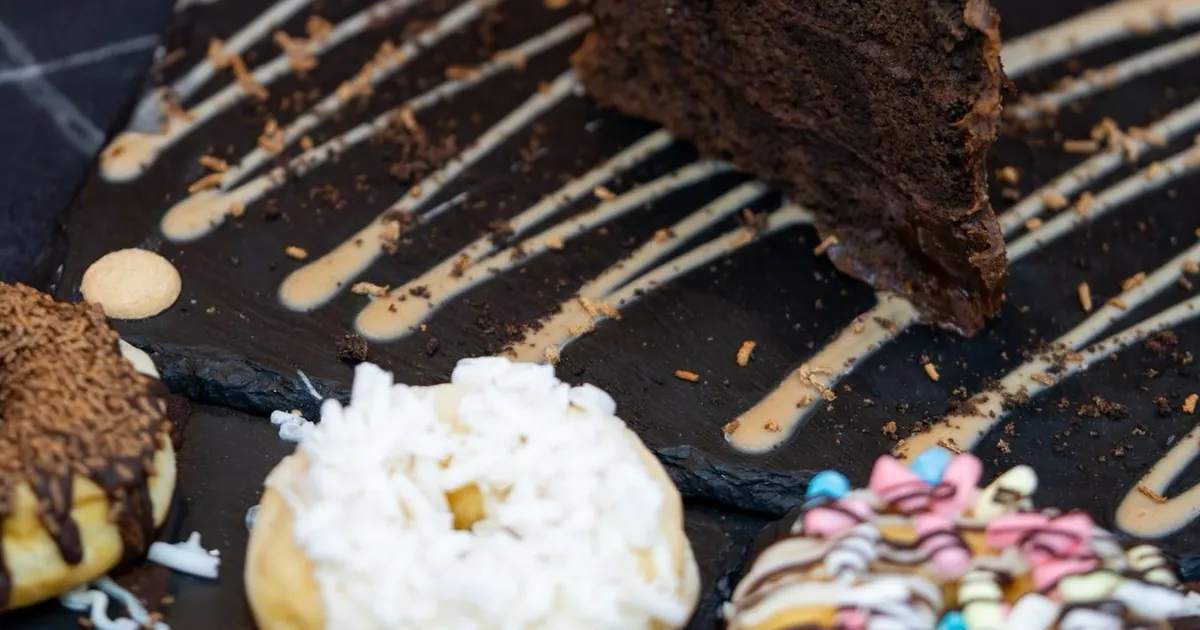 A slice of chocolate cake on black slate streaked with a pale sauce, with coconut, chocolate sprinkle and pastel-iced donuts in the foreground