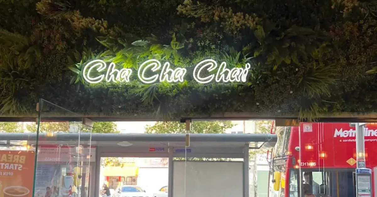 Cha Cha Chai neon sign on a planted wall above the shop door, with a bus shelter and a red bus on Joel Street outside