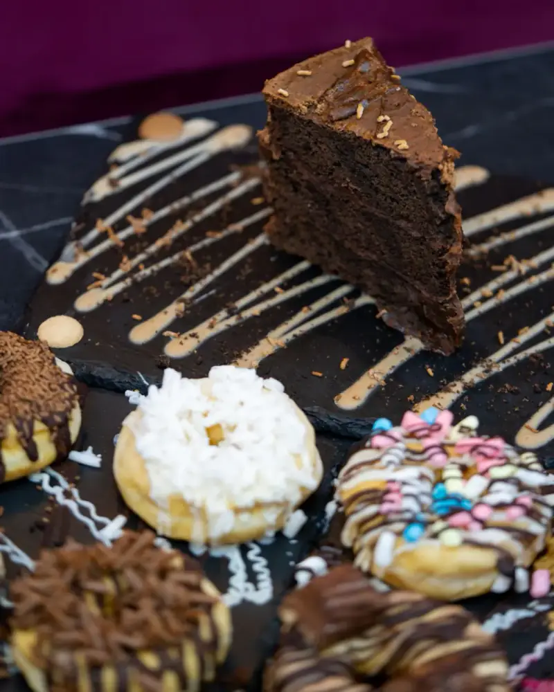 A slice of chocolate cake on a slate drizzled with sauce, with iced and sprinkled donuts around it