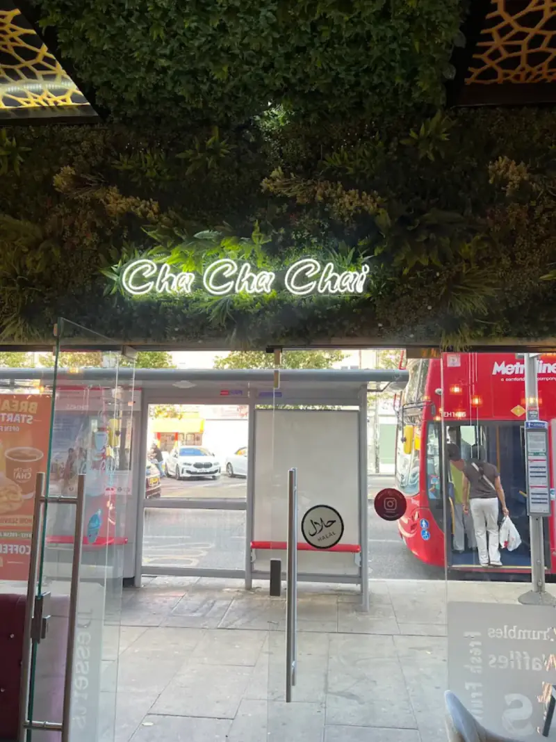 The Cha Cha Chai neon sign over the glass door, with a bus stop and a red bus on Joel Street outside
